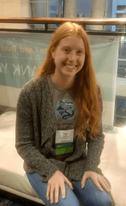 Young woman with long red hair, smiling while seated on a bench in a hotel. She is wearing a conference name tag.