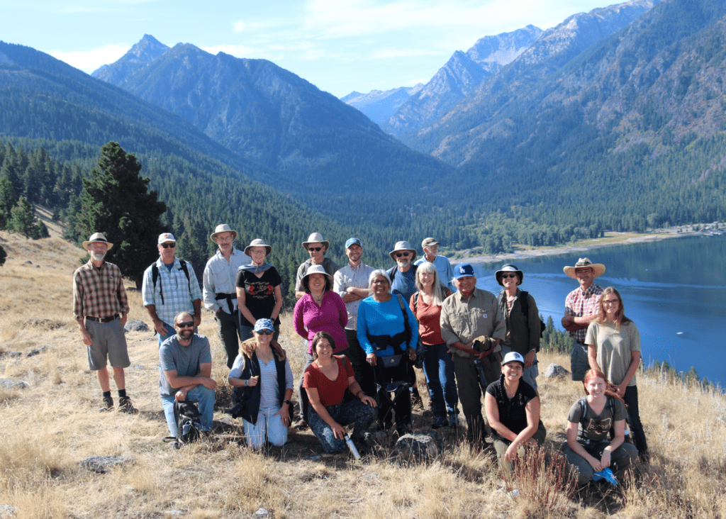 Group of people described in the article atop the East Moraine on a sunny day. There are many smiling faces. The lake and the Wallowa mountain range is present in the background. Dry summer grass is in the foreground.