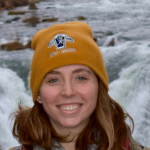 Photo of the author with a yellow beanie standing in front of a waterfall.