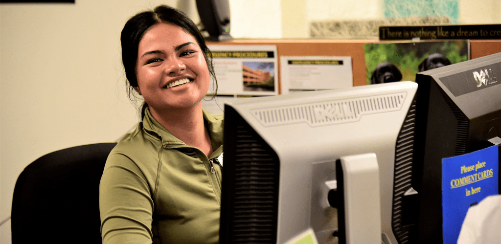 A female college student smiling at the camera while working at a desk and smiling at the camera.