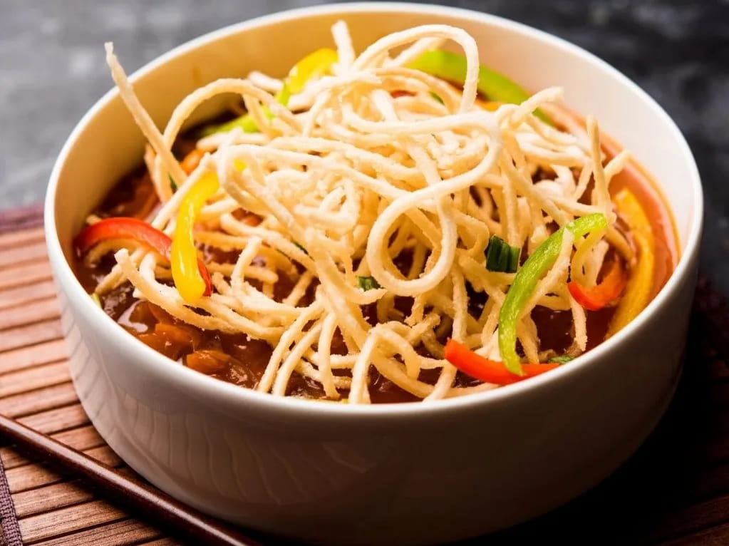 Crispy fried white noodles on top of a bowl of brown sauce chop suey with red and green pepper slivers visible