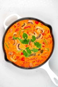 Overhead view of a big pan of creamy tom yum soup: mushrooms, tomatoes, cilantro and basil floating in a creamy, orange soup