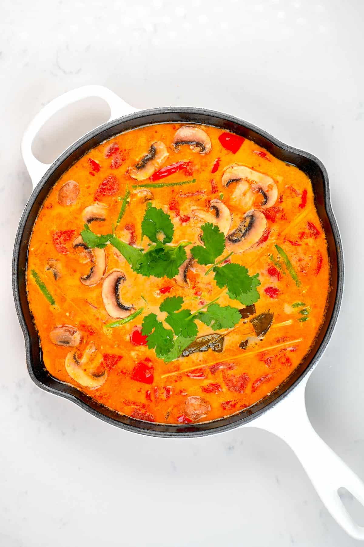 Overhead view of a big pan of creamy tom yum soup: mushrooms, tomatoes, cilantro and basil floating in a creamy, orange soup
