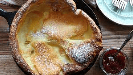 Overhead photo of Dutch Baby with powdered sugar on top; jar of red jam at right