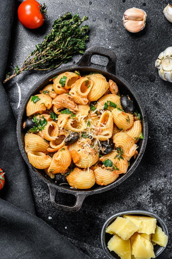 Conchiglie shell pasta with anchovies, tomatoes, garlic and black peppers, in a cast iron dish; tomato, garlic, rosemary and cheese nearby