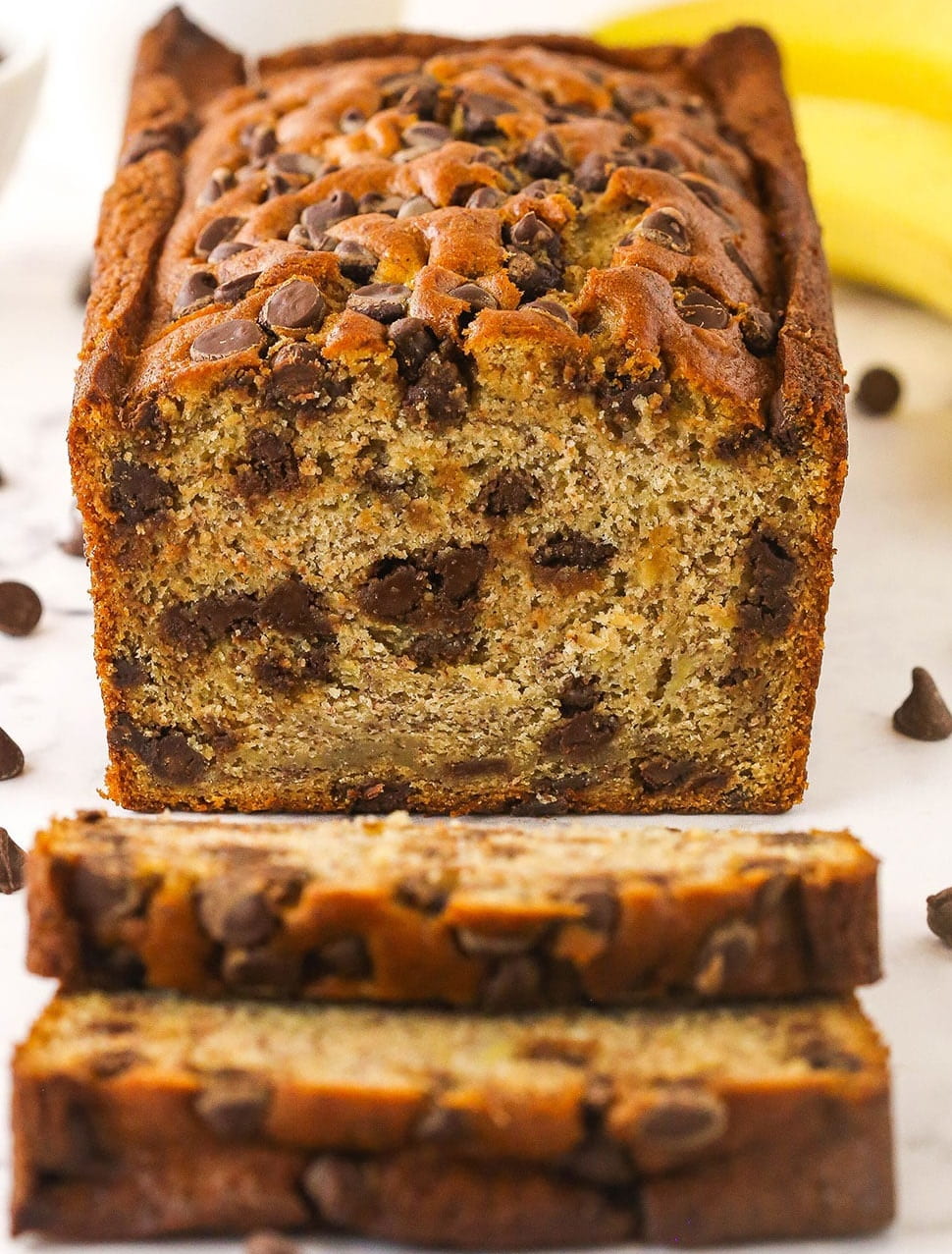 Banana bread loaf with two slices cut from the end and laid flat; chocolate chips visible in the bread and next to the bread; bananas in background; bread on a white table