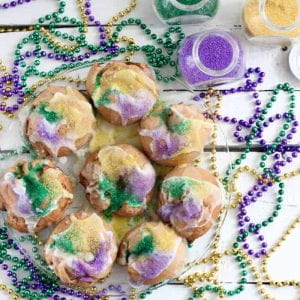 8 small light-brown cakes on a glass plate, decorated with glaze and purple, gold, and green powder. Jars of colored powder and beads (purple, green, and gold) also on the table.