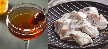 Left: Light brown liquid in a long stem glass with cherries; right: fish pieces in sour cream on a black plate