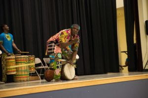 Guinean African master drummer, Alseny Yansane, in traditional clothes, stands smiling over his djembe at the front of a stage. One hand reaches to the drum lying on its side beneath him, the other is outstretched to the crowd.