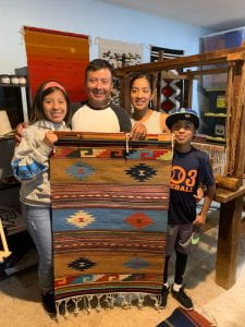Family of teenaged daugther, father, mother, and school-aged son stand behind a traditional woven tapestry of brown, blue stripes with diamond pattern.