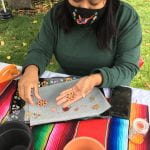 Traditional artist, Diana Gaspar-Sanchez has straight black hair that hnangs below her shoulders and wears a long-sleeved forest green shirt. She sits at a table with a colorfully woven striped tablecloth. Her hand holds an example of her beaded work, the size of a quarter, over a tray of several more examples. shows Zapotec Beading