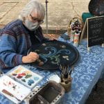 The image shows traditional artist, Patti Jo Meshnik, with mid-length grey hair and black frame glasses painting floral patterns on a round black plate about 18" diameter. She demonstrates this Norwegian Rosemåling at a table with blue table cloth at Whiteaker Community Market in Eugene, Oregon, Oct 3, 2021