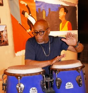 Jardin Kazaar is a black man with black glasses and a small white beard on his chin. He is playing on the conga drums.