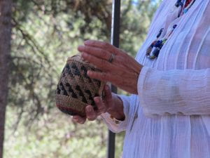 Indigenous woman's hands hold a small, cup-shaped basket of natural brown and black weave.