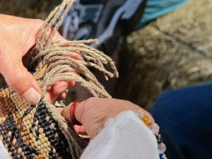 Two Indigenous hands hold a basket mid-creation with cordage. An amber ring on her right thumb and her left fingers are nestled among the weavers.