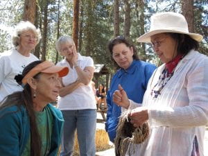 Pat has shoulder-length black hair held back by a white, brimmed sunhat, and tinted eyeglasses. She stands in the foreground, circled by four women watching attentively. Hanging in Pat's left hand is a basket in-progress and her right hand points to the strands. Tall evergreen trees tower in the background.