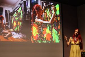 Woman with long red hair and white dress stands on stage before a projected image of herself painting.