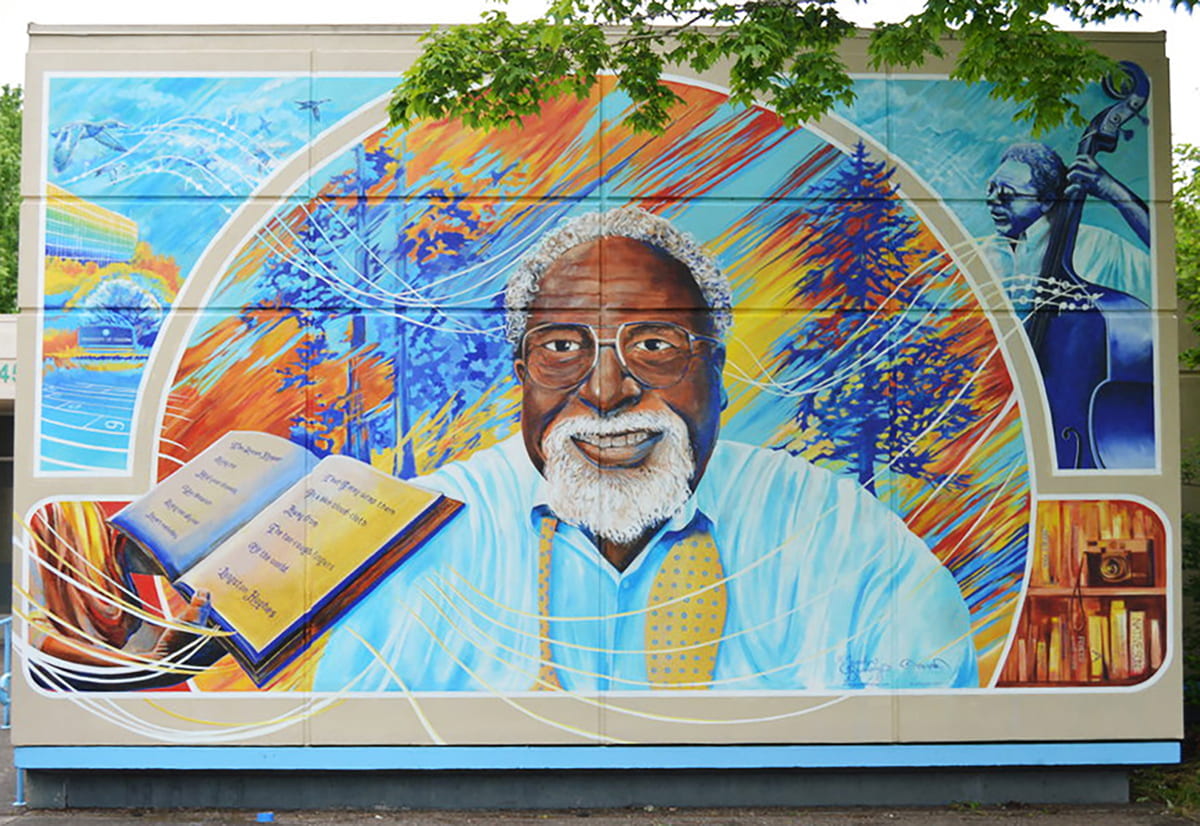 Colorfully painted mural of Ed Coleman smiling with a blue background depicting trees and nature. Surrounding him are illustrations of books and musicians.