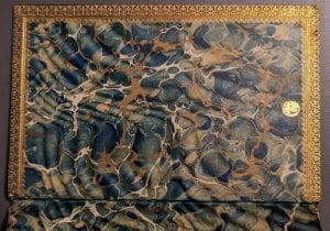 Inner cover of a book with pages marbled in brown, green and blue and edged in gold. A small gold seal is on the right.