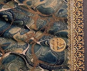 Inner cover of a book with pages marbled in brown, green and blue and edged in gold. A small gold seal is on the right.
