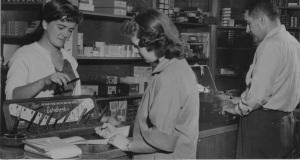 Black and white photo of three people in a store. One, who presents as a woman, stands behind a counter and holds a pen attached to a pen display. A person who presents as a woman faces her, holding another pen above a piece of paper. The third person, who presents as a man, stands a typewriter that sits on the counter. In the background, shelves are filled with boxes of varied sizes. 