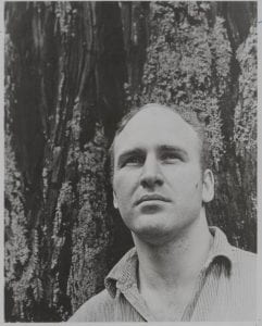 Black and white portrait of Kesey, who stands in front of a tree. He is balding, has light skin tone and a collared shirt. 