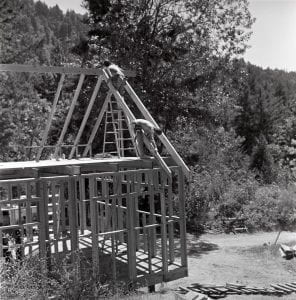 Two topless figures frame a wooden structure in the woods. 