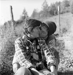 Black and white photo of two female-presenting people, locked in an embrace. The person in front has short hair and wears a plaid shirt, with a camera hanging from their neck. The other person sits behind them, wearing a beanie hat, their arms wrapped around the first person. 
