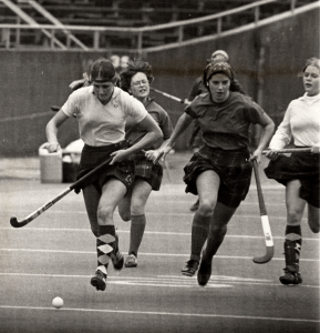 Black and white photo of five women playing field hockey. They run down a field wearing tee shirts, knee socks and skirts.