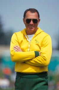 A person with light skin and short dark hair wears sunglasses and a yellow jacket, standing smiling with their arms crossed on their chest. 