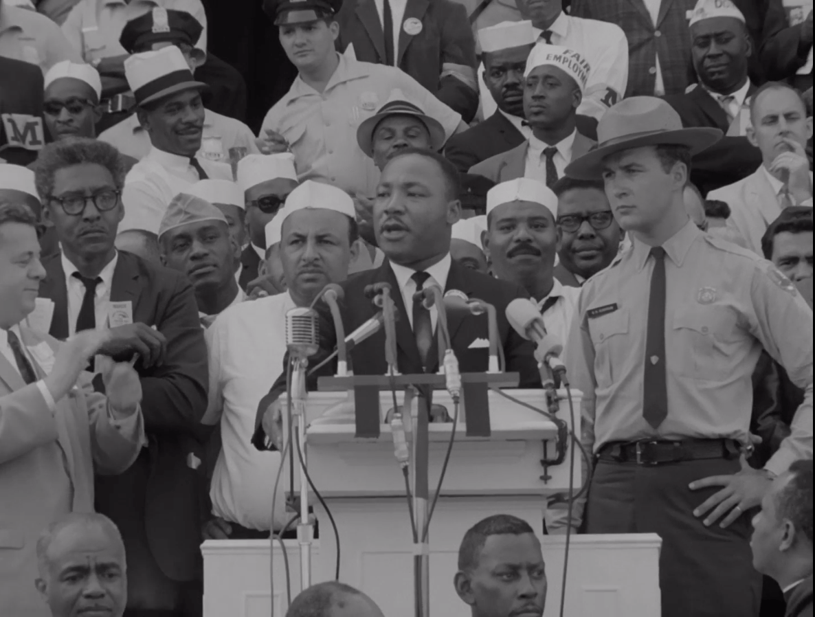 Martin Luther King, Jr., speaks at a podium. On his left is Bayard Rustin, and on his right is a police officer.