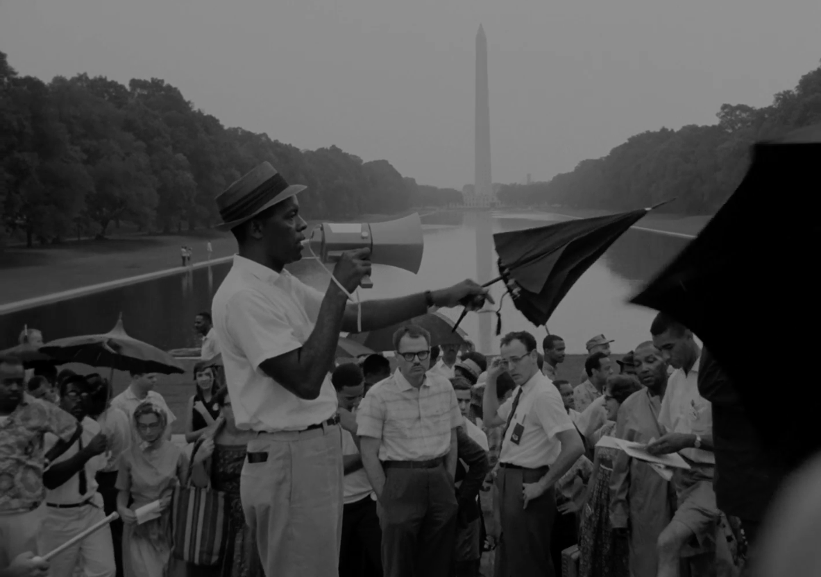 A man holding a megaphone gestures with an umbrella in front of a crowd of people.