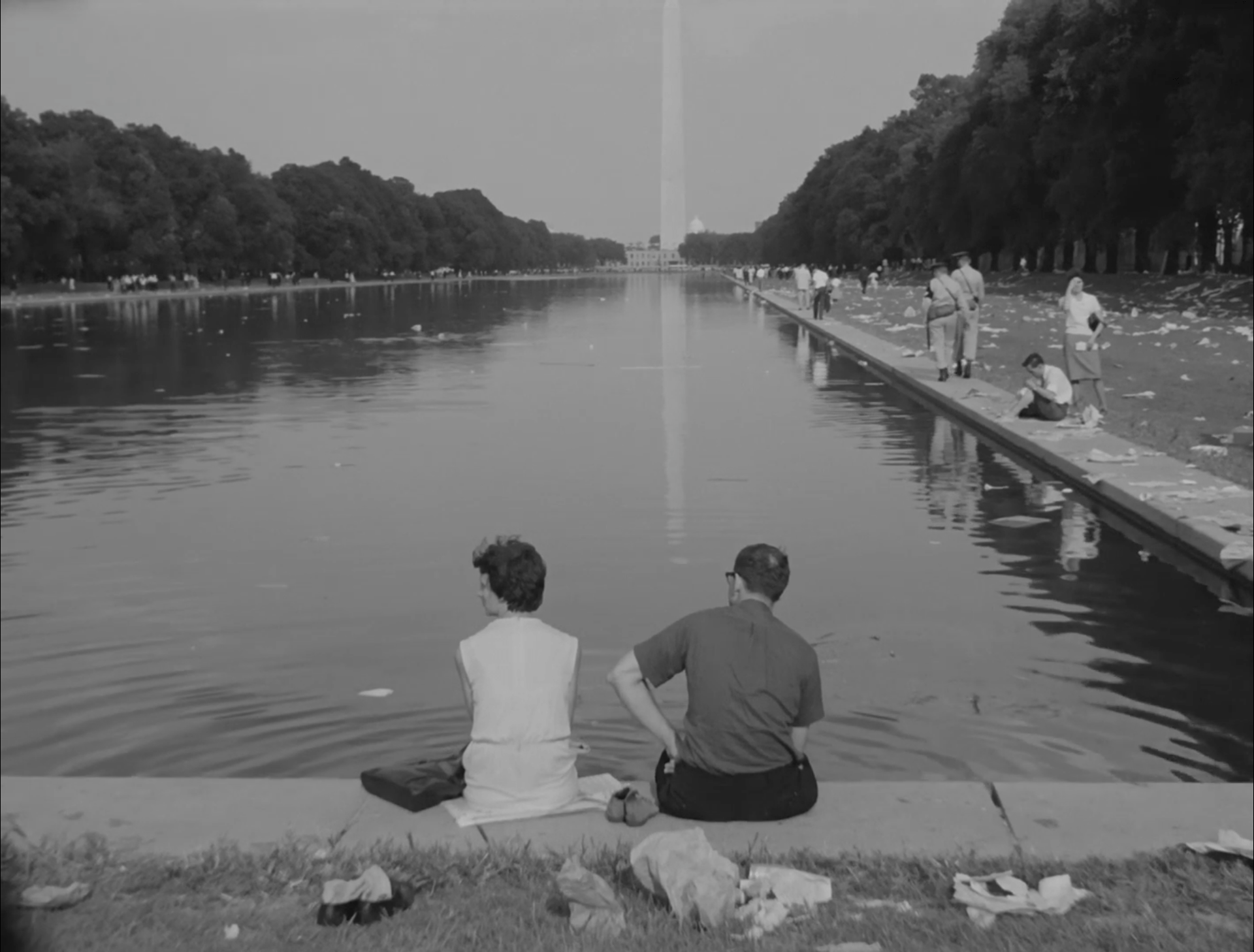 People sit on the edge of the reflecting pool on the National Mall (Washington, DC), facing the Washington Monument. There is litter on the ground left after the March on Washington.