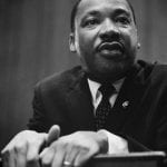 A portrait of the Reverend Doctor Martin Luther King Jr., clasping the back of a chair.